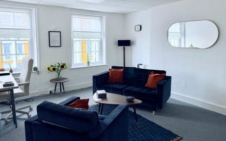 A modern, light-filled office with dark sofas, a coffee table, and a workspace.